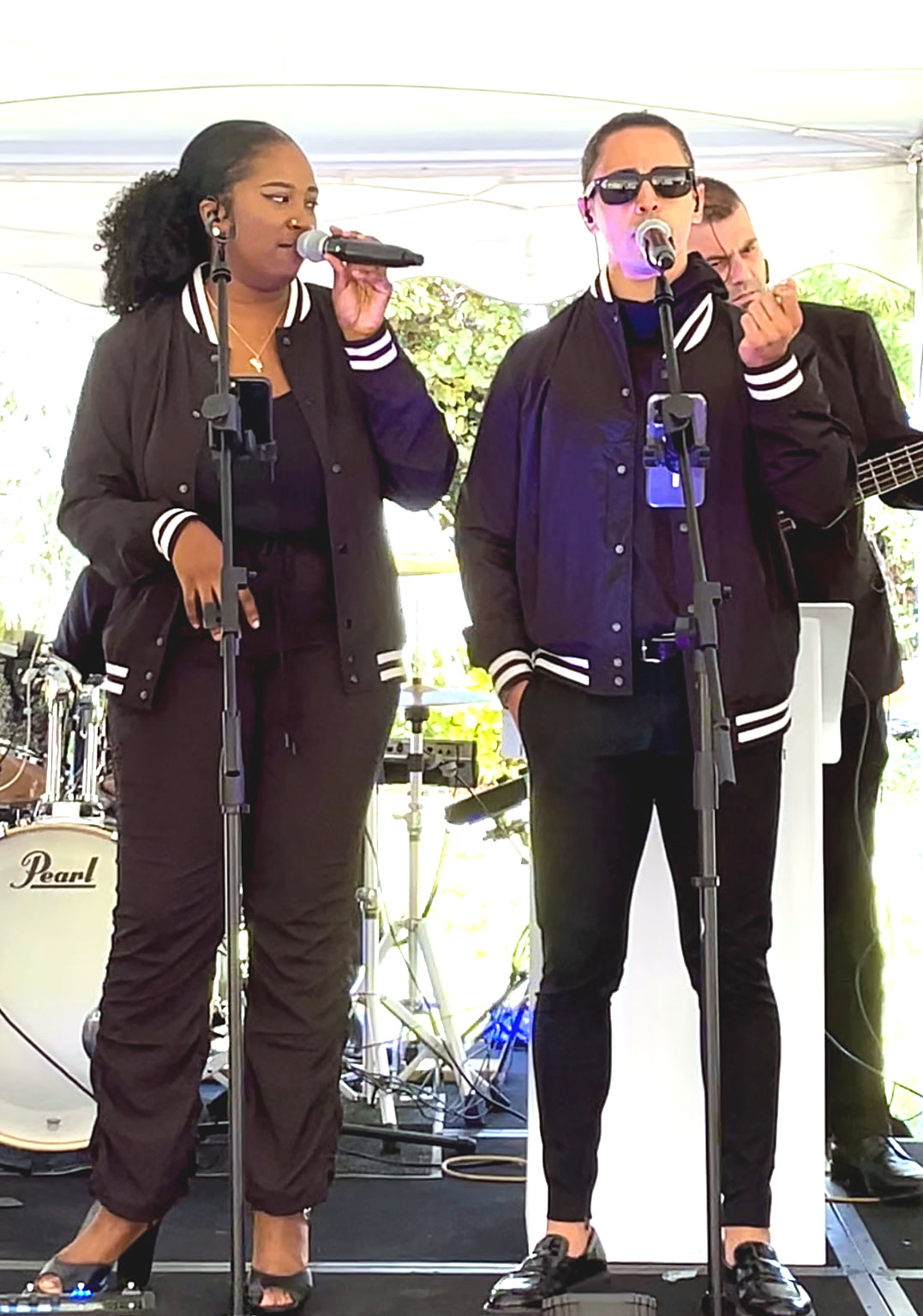 Two Gordon Dukes Music Company vocalists in varsity jackets singing under a tent with bass and drums behind them