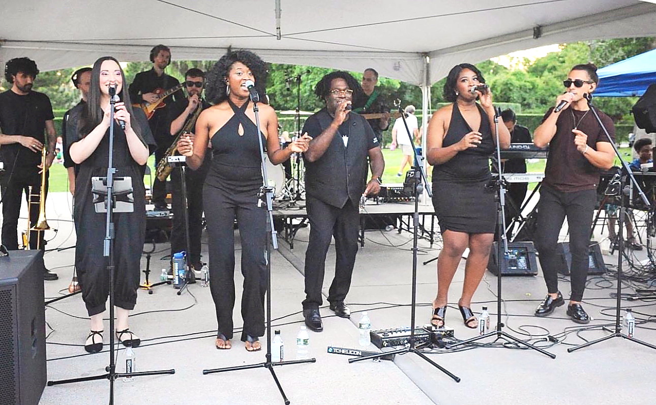 Five Gordon Dukes Music Company vocalists singing under a tent with saxophone, trombone, guitar and keyboards behind them