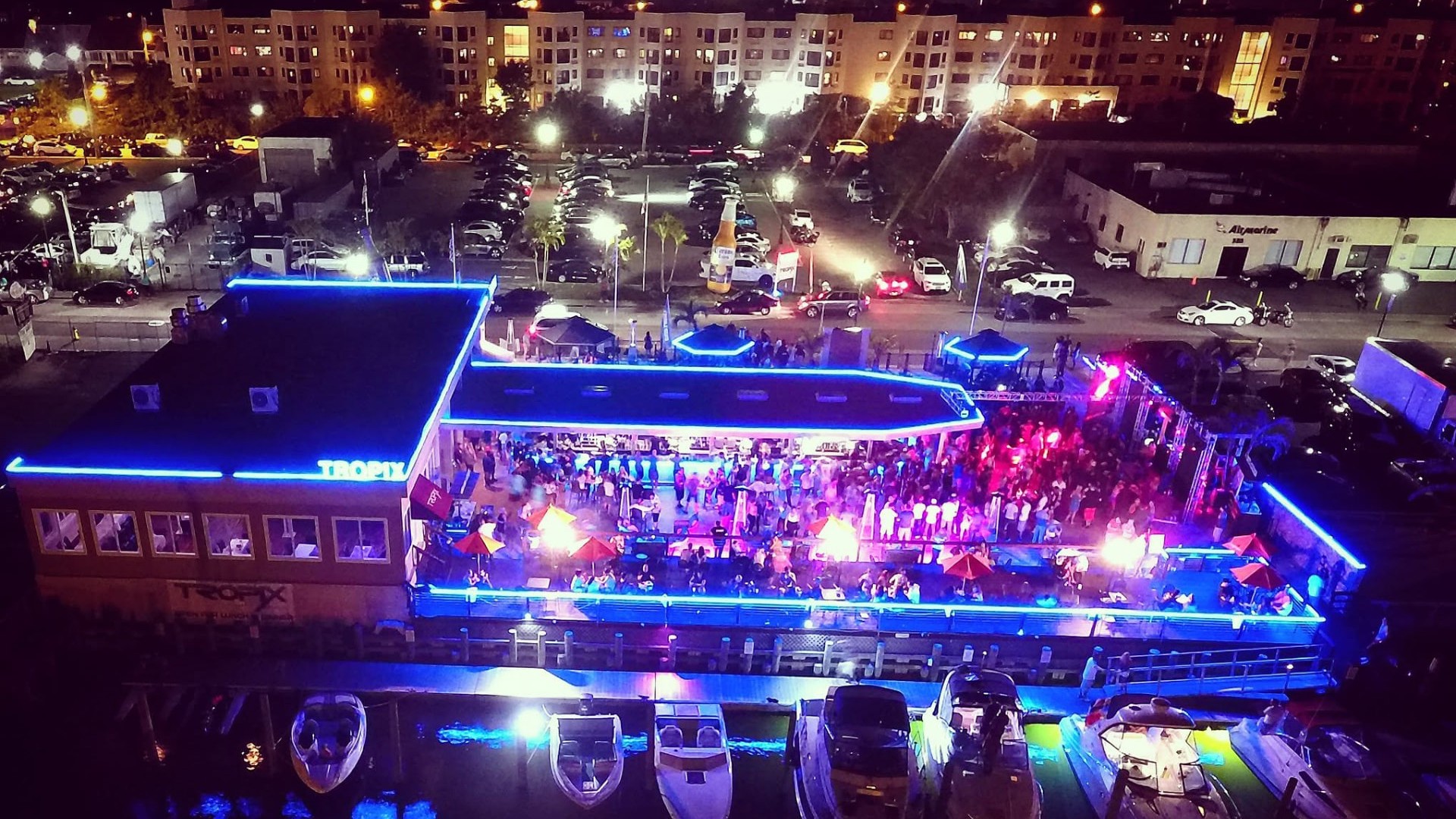 An illuminated waterfront venue at night with a full crowd on the open-air deck and boats moored along the marina — Gordon Dukes Music Company