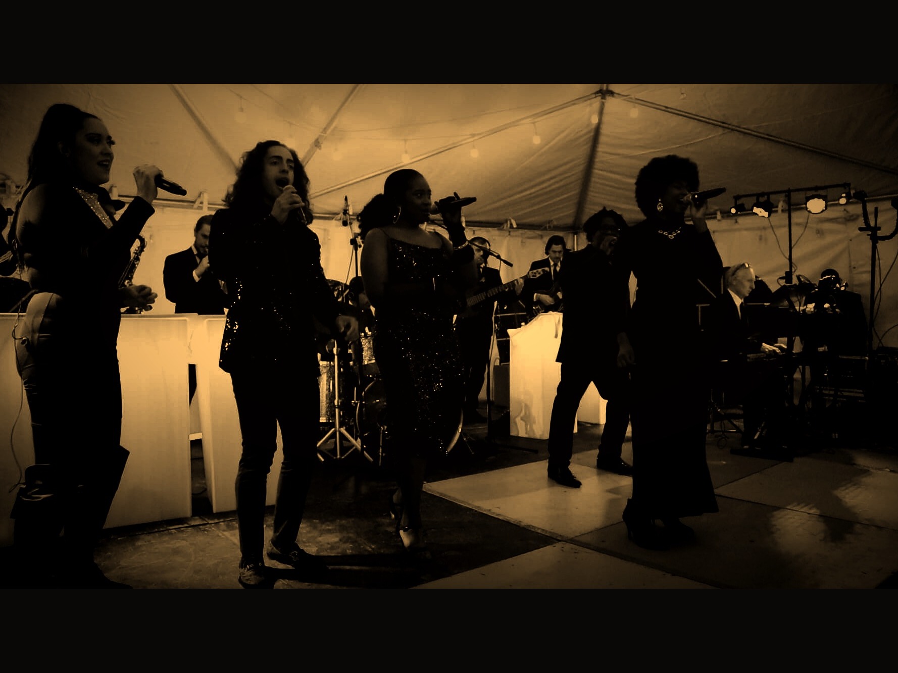 Four vocalists at the front of a full live band mid-performance on a concert stage — Gordon Dukes Music Company