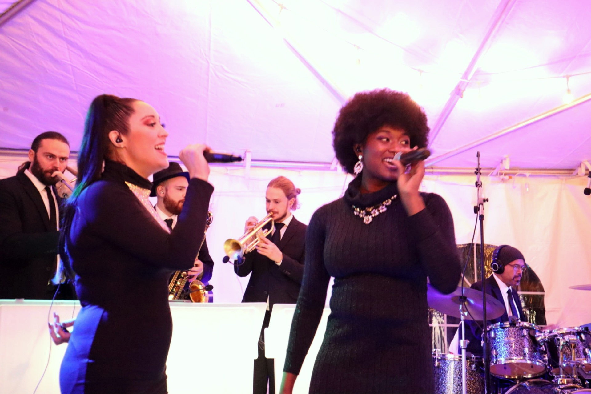 Two Gordon Dukes Music Company lead vocalists singing on a tented stage with the horn section and drummer behind them