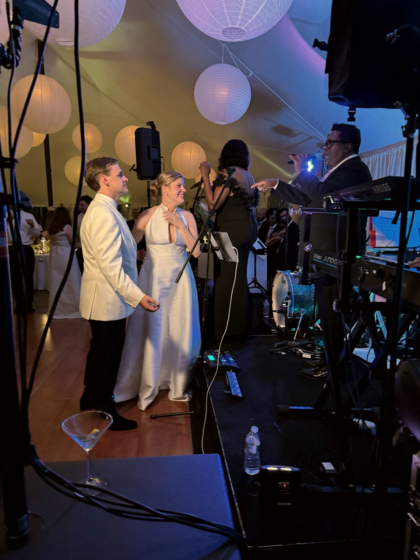 Gordon Dukes sings from the bandstand and gestures toward the newlyweds standing beside the stage under a lantern-lit tent