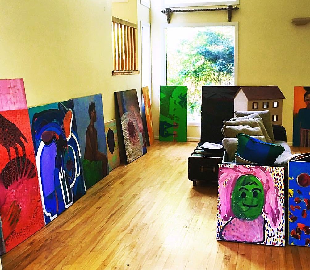 Finished paintings lined along the walls of a working artist's studio in tall natural daylight — Imagine If Foundation, Gordon Dukes Music Company