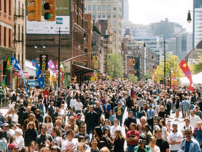 A dense crowd filling a closed New York City avenue during a large public street event, with banners, storefronts and city buildings framing the block