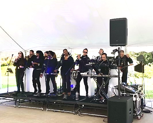 The full Gordon Dukes Music Company ensemble — vocalists, rhythm section and keyboards — performing under a white tent at an outdoor evening performance