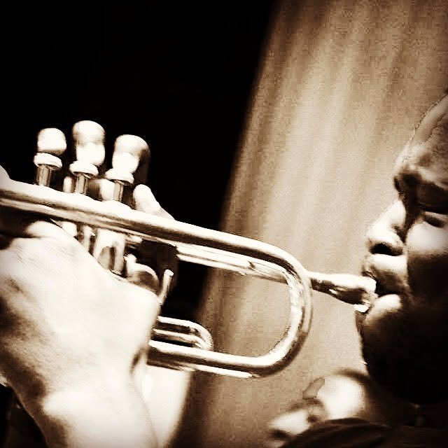 Donovan Dukes playing trumpet in low stage light, warm sepia tone